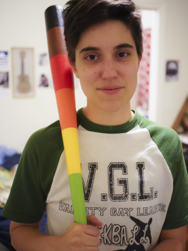 Photo of a queer person with short hair holding a rainbow colored baseball bat.
